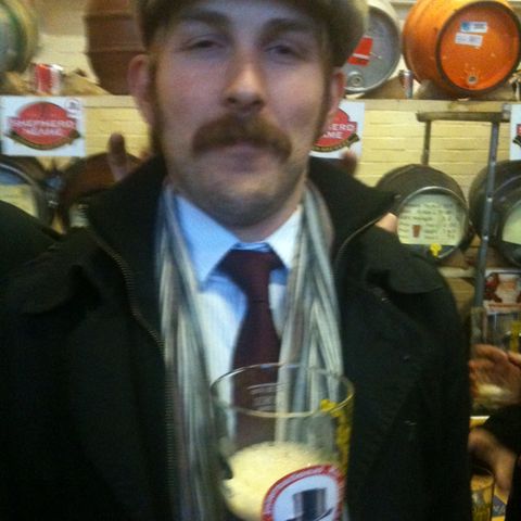 A man with a fine moustache in a flat cap and tie, holding up a pint at a beer festival