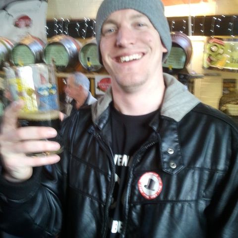A grinning man in a grey beanie raises a glass of beer