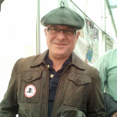 A man in a tweed flat cap and leather jacket with a glass of ale