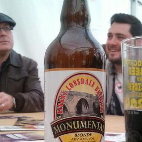 Two men at a festival table, one in a flat cap, behind a bottle of ale