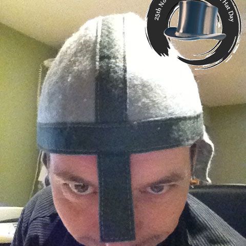A man in a grey knitted knight's-helmet hat, holding up the 2012 logo